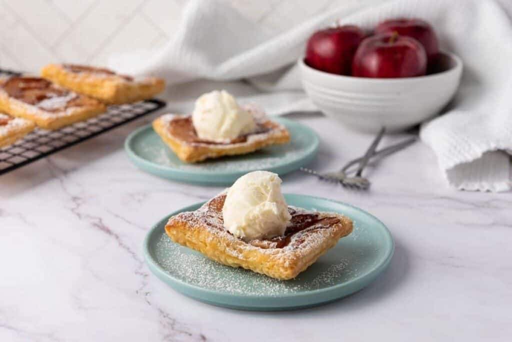 Two square pastries topped with a scoop of vanilla ice cream are served on blue plates, with a bowl of red apples and more pastries in the background.