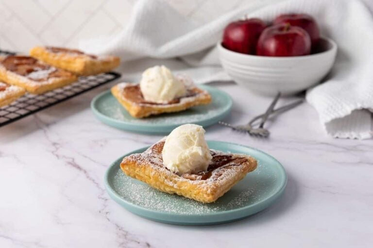 Two square pastries topped with a scoop of vanilla ice cream are served on blue plates, with a bowl of red apples and more pastries in the background.