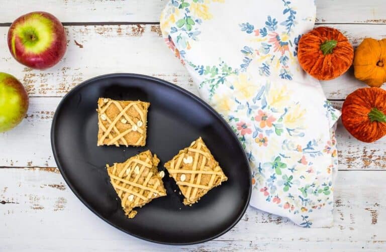 Three Brown Butter Apple Blondies on a black oval plate, with apples, small pumpkins, and a floral napkin on a white wooden surface.
