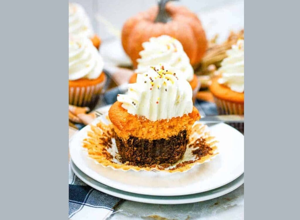 A cupcake with orange pumpkin and dark chocolate layers, topped with swirled white frosting and sprinkles, sits on a white plate with its wrapper partially peeled back.