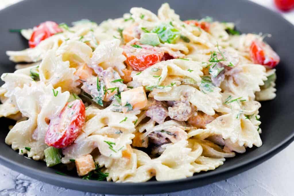 A black plate filled with bowtie pasta mixed with creamy sauce, diced vegetables, cherry tomatoes, herbs, and small chunks of meat.