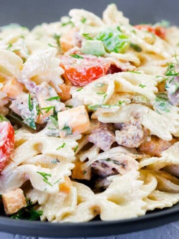A black plate filled with bowtie pasta mixed with creamy sauce, diced vegetables, cherry tomatoes, herbs, and small chunks of meat.
