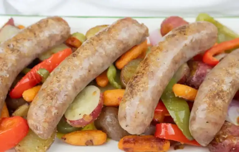 Four cooked sausages served on a bed of roasted vegetables including red potatoes, carrots, red bell peppers, and green bell peppers.