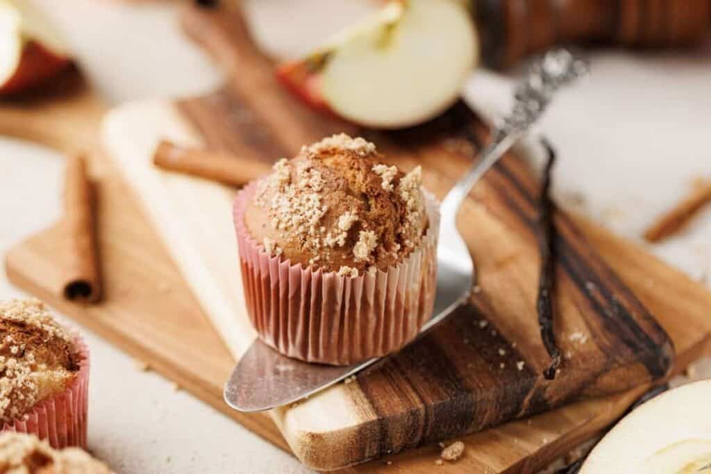 A muffin with crumb topping sits on a butter knife atop a wooden cutting board, surrounded by apple slices, a cinnamon stick, and a vanilla bean pod.