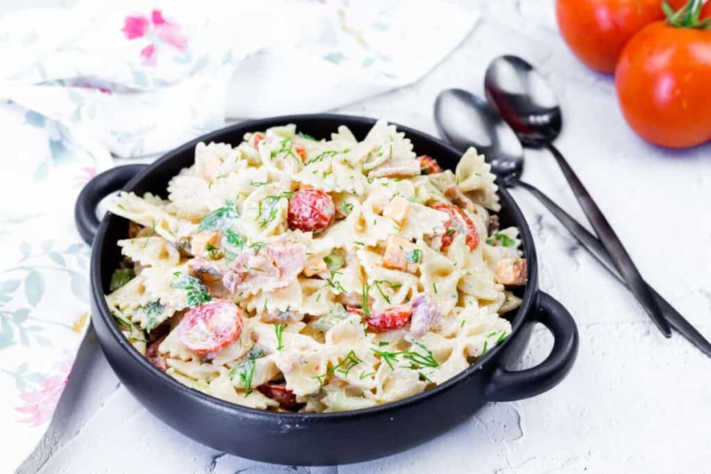 A black bowl filled with creamy bowtie pasta salad with chopped vegetables and herbs, placed on a white surface next to fresh tomatoes and utensils.