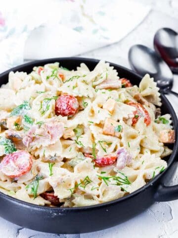 A black bowl filled with creamy bowtie pasta salad with chopped vegetables and herbs, placed on a white surface next to fresh tomatoes and utensils.