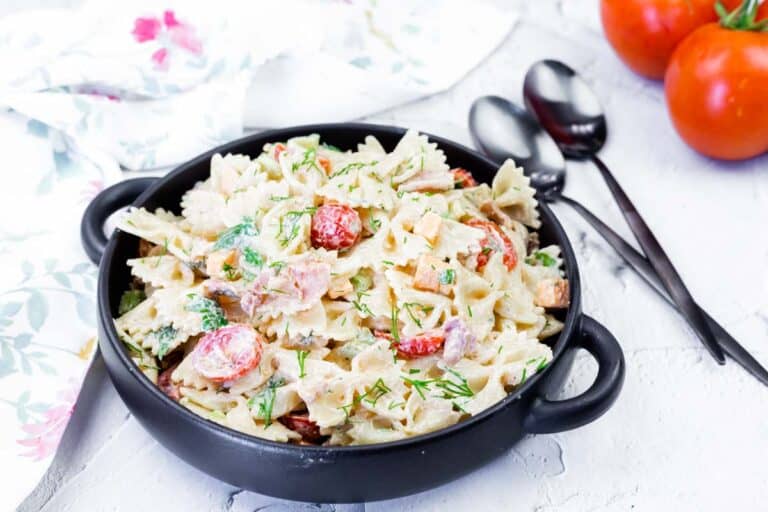 A black bowl filled with creamy bowtie pasta salad with chopped vegetables and herbs, placed on a white surface next to fresh tomatoes and utensils.