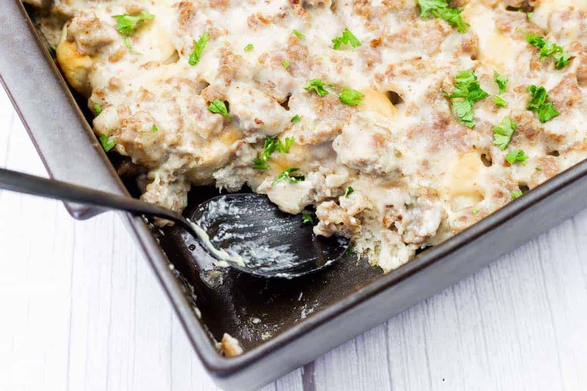 A baking dish of sausage and biscuit casserole topped with creamy gravy and chopped parsley, with a serving spoon partially in the dish.