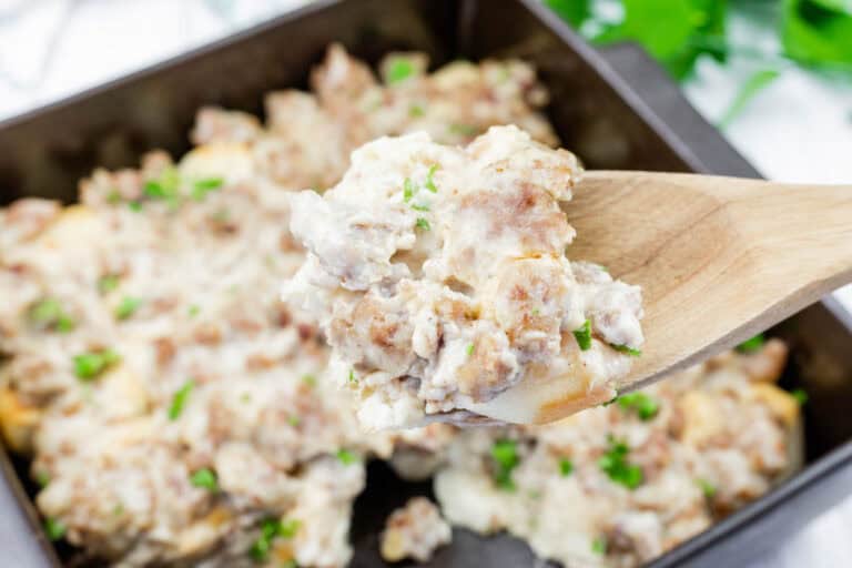 A wooden spoon holds a serving of biscuits and gravy with sausage, over a baking dish filled with more of the same dish, garnished with chopped herbs.