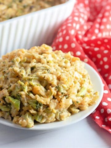A plate of Easy Cheesy Broccoli Rice Casserole sits on a white surface next to a red and white polka dot cloth, with a baking dish in the background.