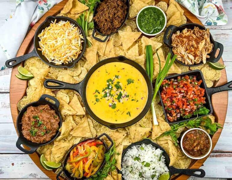 A round platter with tortilla chips, queso dip in the center, and small bowls of shredded cheese, meats, salsa, beans, rice, fajita vegetables, and green sauce arranged around the edge.