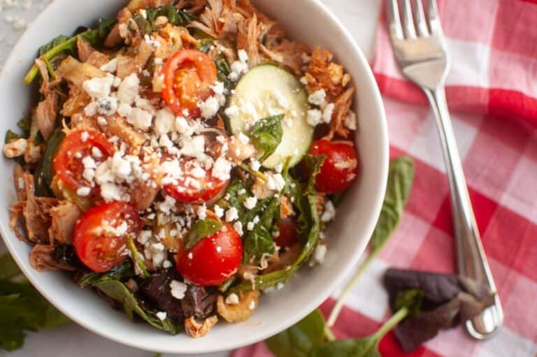 A bowl of salad with shredded meat, cherry tomatoes, spinach, cucumber slices, and crumbled cheese, next to a fork and a red-checkered napkin.
