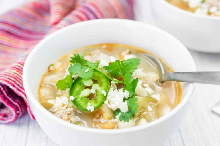 A bowl of soup garnished with sliced jalapeño, cilantro, and crumbled cheese, with a spoon resting inside and a colorful cloth napkin nearby.