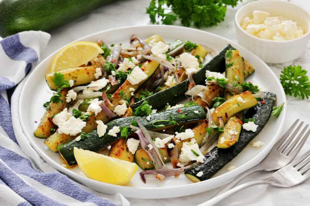 A plate of saut&eacute;ed zucchini with onions, topped with crumbled cheese, parsley, and lemon wedges, with a fork and napkin on the side.