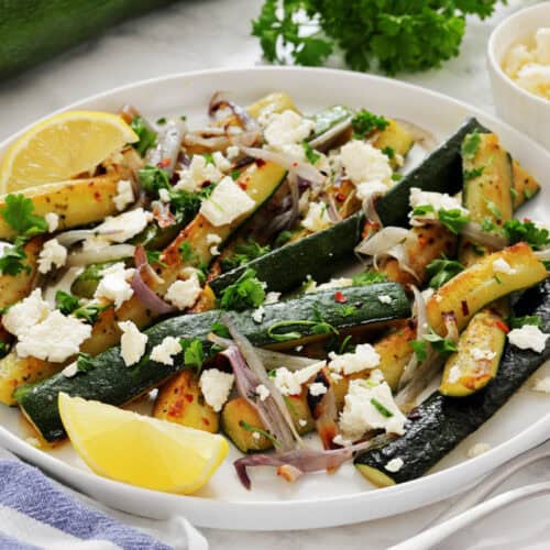 A plate of saut&eacute;ed zucchini with onions, topped with crumbled cheese, parsley, and lemon wedges.