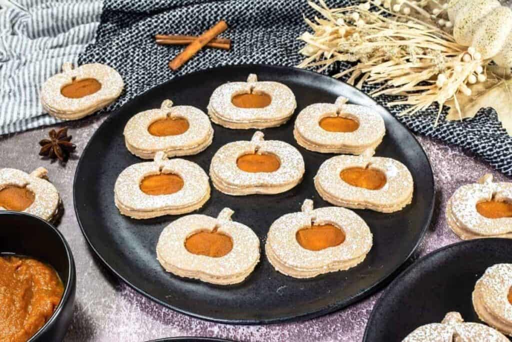 Round pumpkin-shaped sandwich cookies with orange filling and powdered sugar on a black plate, surrounded by autumn-themed decorations and ingredients.