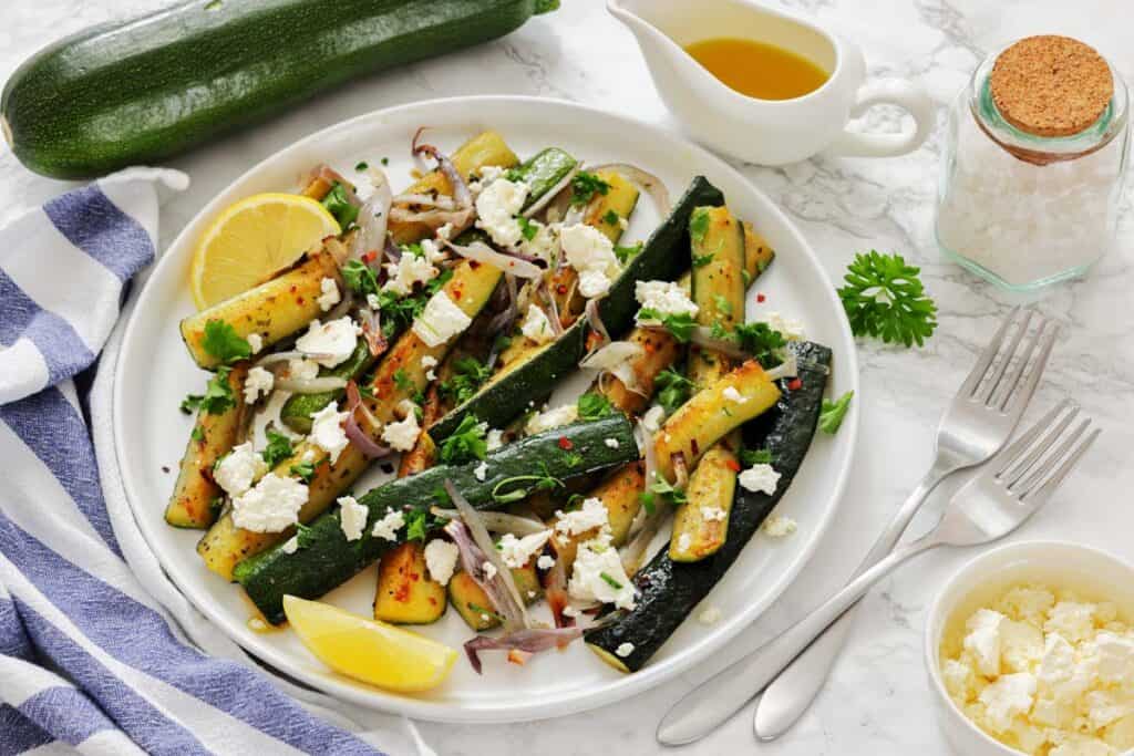 A plate of grilled zucchini with crumbled feta, red onion, herbs, and lemon wedges, accompanied by olive oil and salt, sits on a table with cutlery and a napkin.