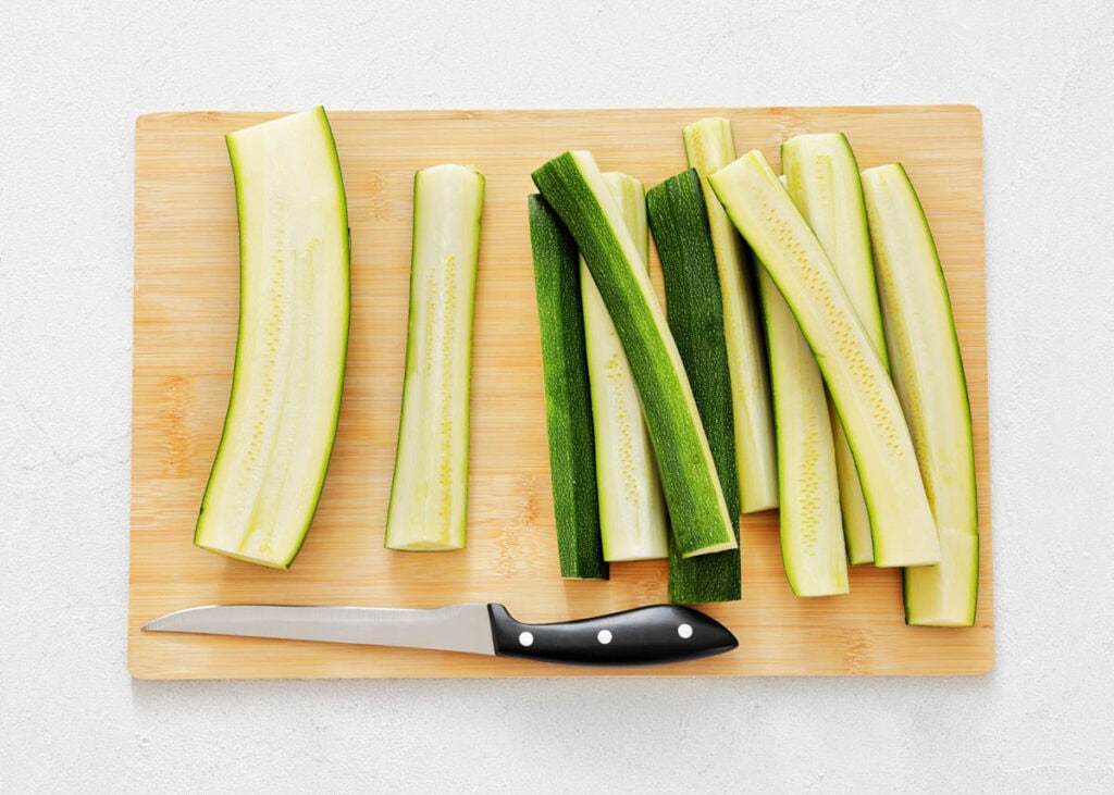 Sliced zucchini sticks and a black-handled knife are arranged on a wooden cutting board.