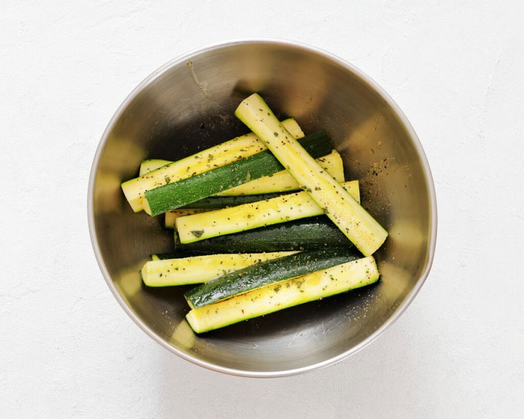Metal bowl containing raw zucchini sticks seasoned with herbs, salt, and pepper, placed on a white surface.