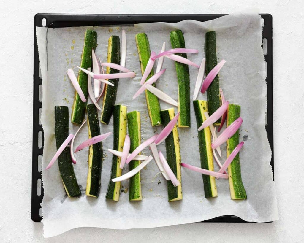 Baking tray lined with parchment paper holding zucchini spears and sliced shallots, drizzled with oil, ready for roasting.