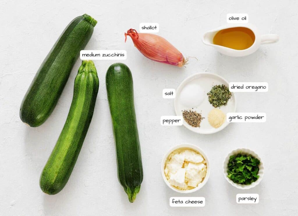 Three medium zucchinis, a shallot, olive oil, salt, pepper, dried oregano, garlic powder, feta cheese, and chopped parsley are arranged on a white surface.