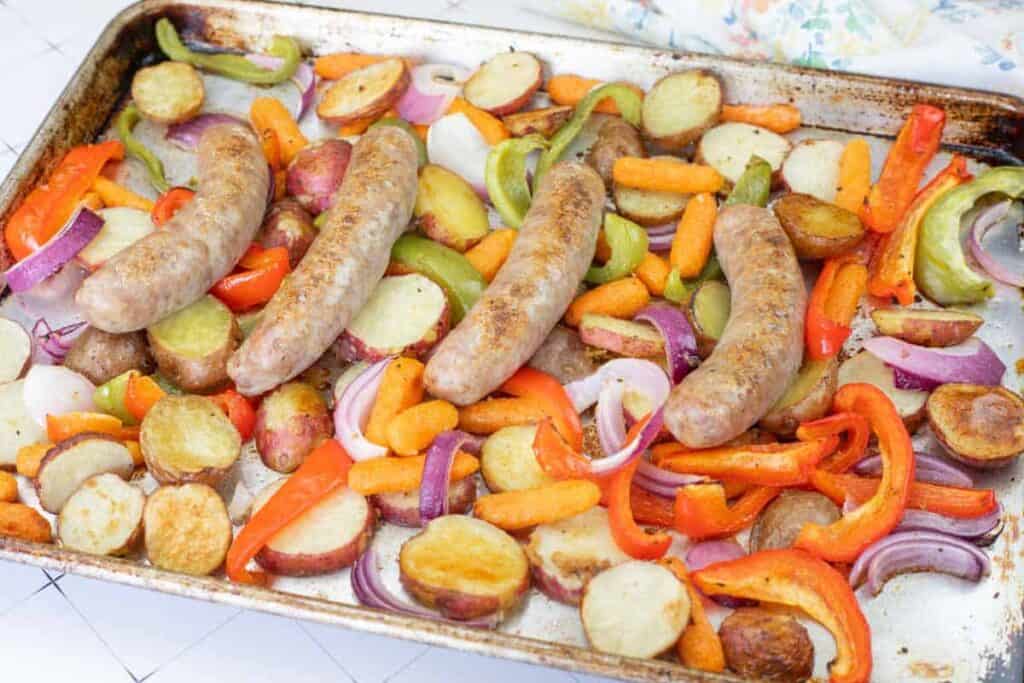 Sheet pan with cooked sausages, sliced potatoes, carrots, red and green bell peppers, and red onions.