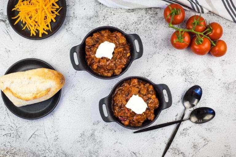 Two small bowls of chili topped with sour cream, a plate of shredded cheese, a baguette, fresh tomatoes on the vine, and two spoons on a light surface.