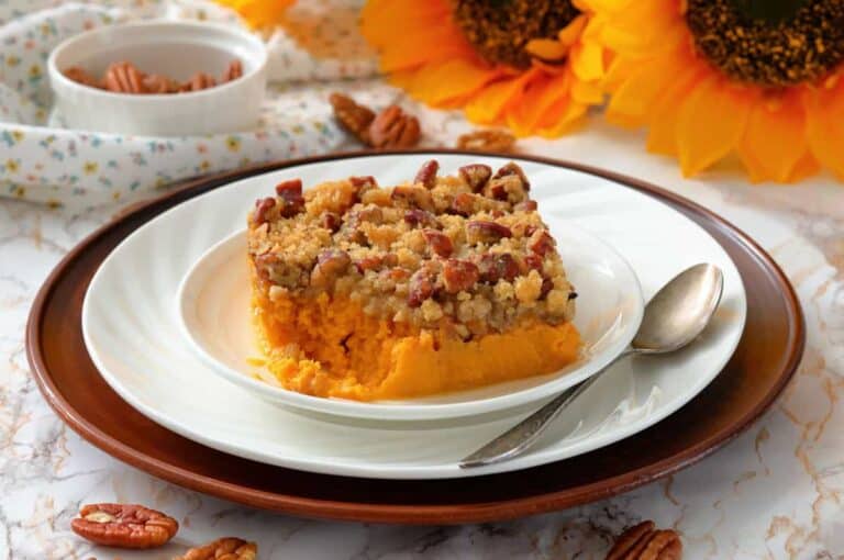A slice of sweet potato casserole topped with a pecan crumble sits on a white plate with a spoon, surrounded by pecans and sunflowers.