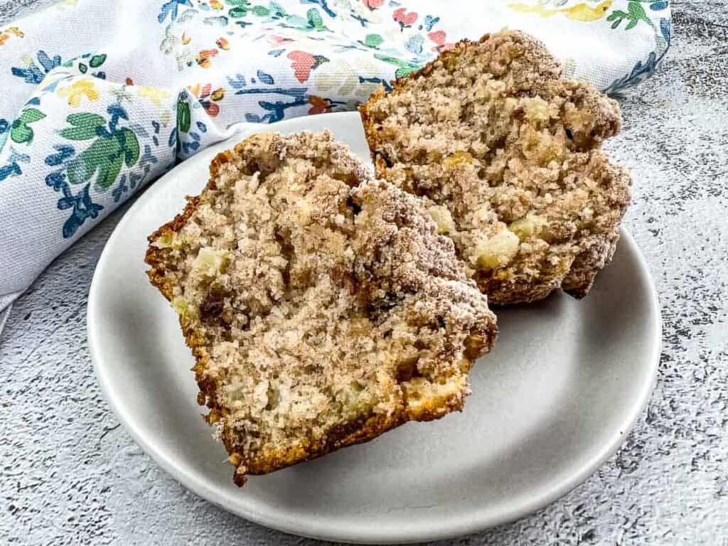 Two halves of a crumb-topped muffin with visible apple pieces are on a white plate, set on a light textured surface beside a floral-patterned cloth.