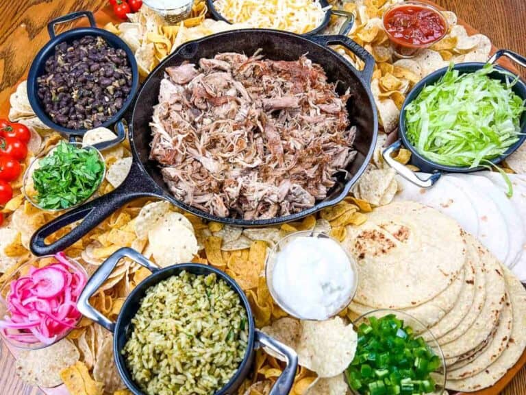 A serving platter with shredded pork in a cast iron skillet, surrounded by tortilla chips, tortillas, rice, beans, lettuce, salsa, sour cream, cheese, and various toppings.