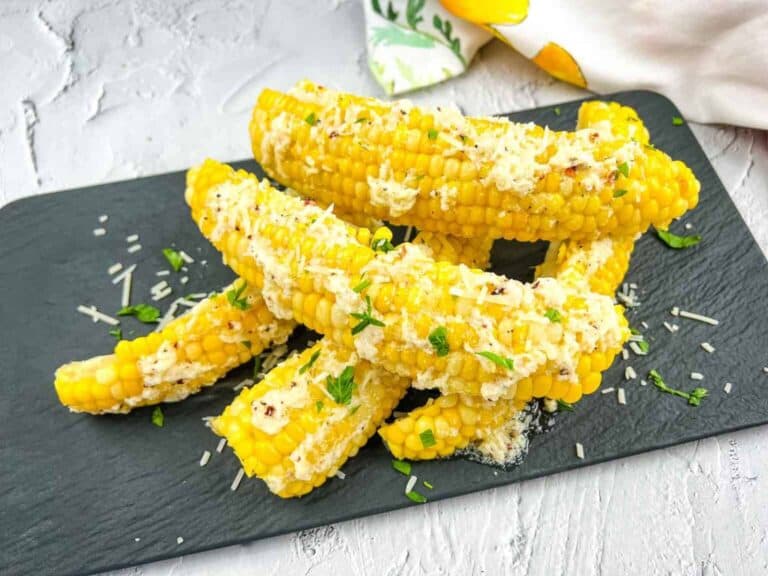 Six pieces of corn on the cob topped with grated cheese and chopped herbs are arranged on a black slate serving tray.