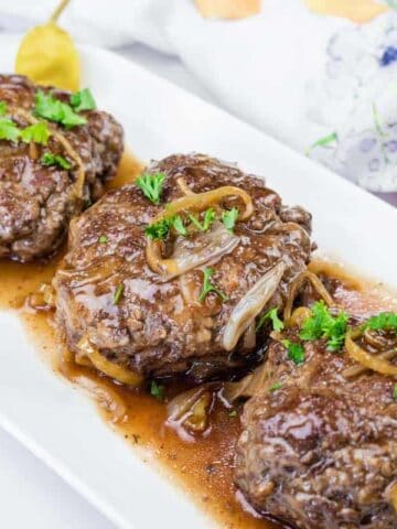 Three Salisbury steak patties topped with onions and gravy, garnished with parsley, served on a white rectangular plate.