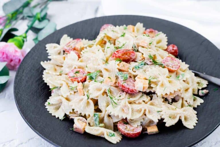 A black plate filled with farfalle pasta mixed with a creamy sauce, cherry tomatoes, chopped vegetables, and garnished with fresh herbs.