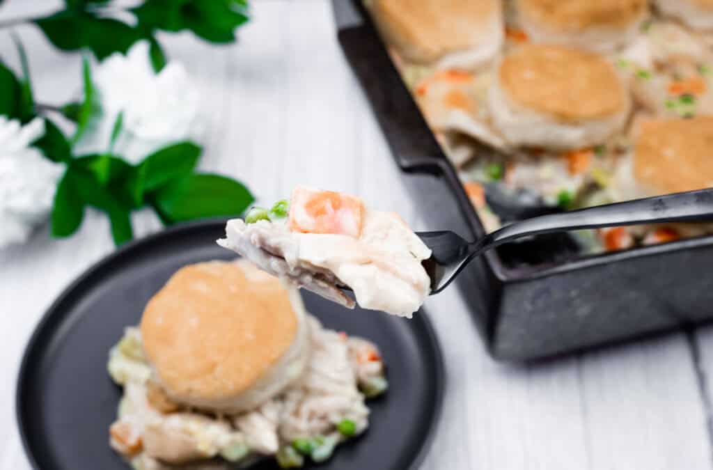A fork holds a bite of biscuit-topped chicken pot pie with vegetables, with a serving on a black plate and the baking dish in the background.