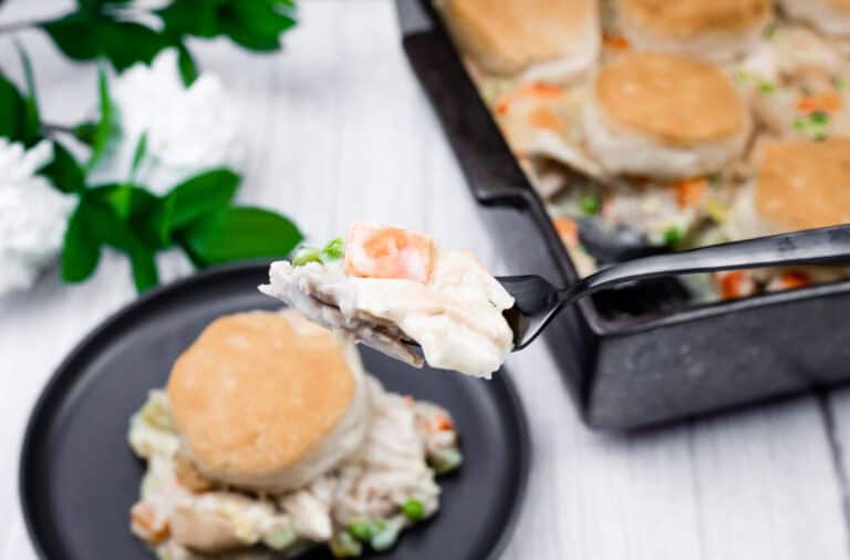 A fork holds a bite of biscuit-topped chicken pot pie with vegetables, with a serving on a black plate and the baking dish in the background.