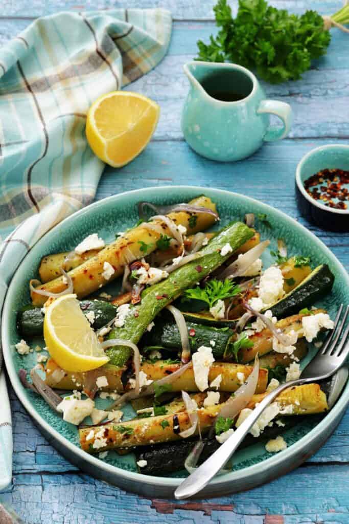 A plate of grilled zucchini and squash with onions, feta cheese, and herbs, garnished with lemon wedges. A fork, cloth napkin, sauce, and parsley are placed nearby on a blue wooden surface.
