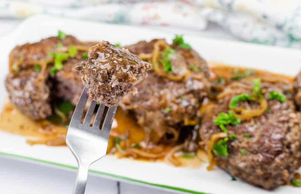 A close-up of a fork holding a bite of Salisbury steak with gravy and onions, with more steak pieces and sauce garnished with parsley in the background.