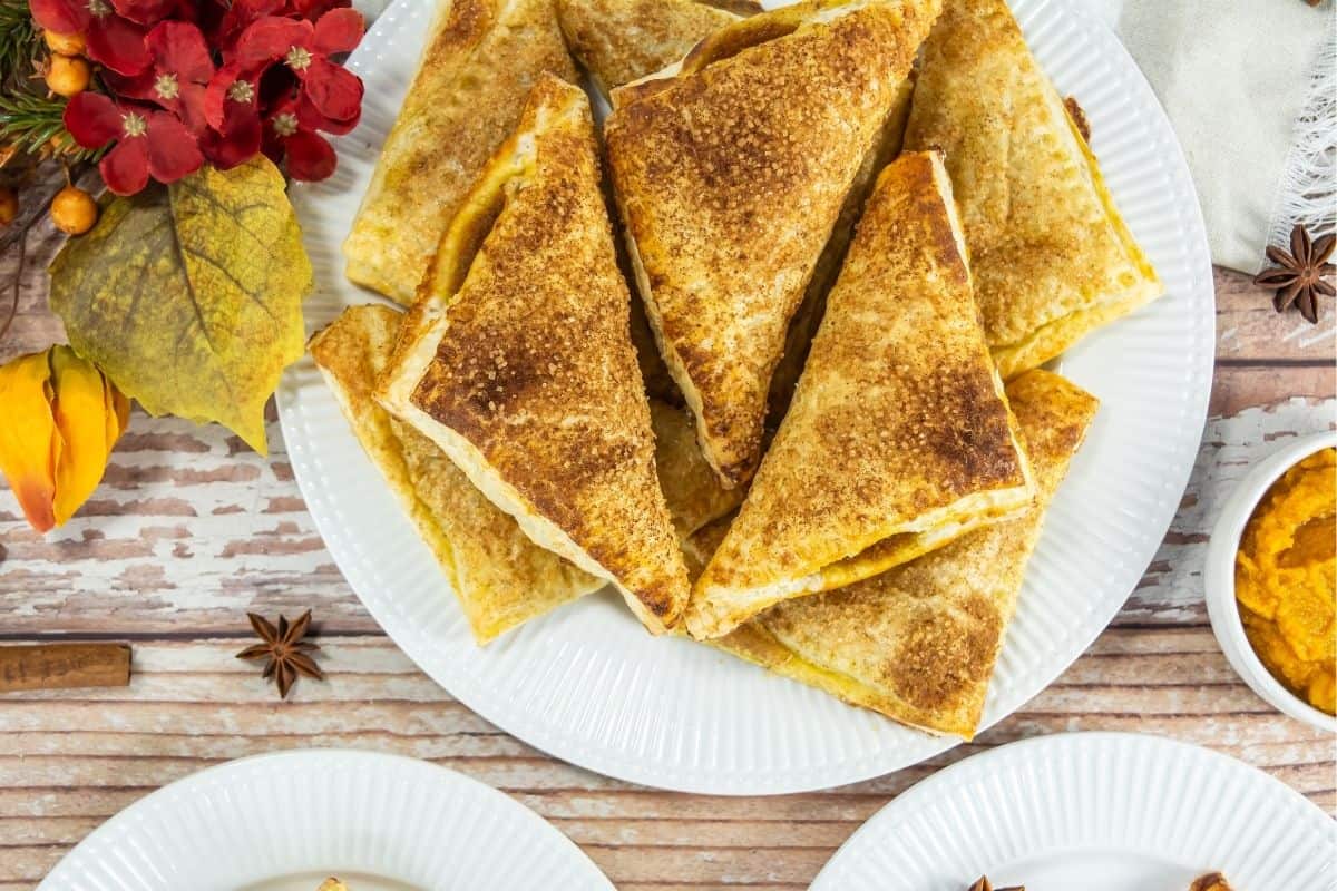A plate of Pumpkin Turnovers arranged in a stack, with autumn-themed decorations and a small bowl of orange spread nearby.
