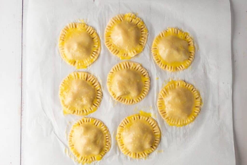 Nine round, uncooked hand pies with crimped edges and egg wash are arranged on a sheet of parchment paper.