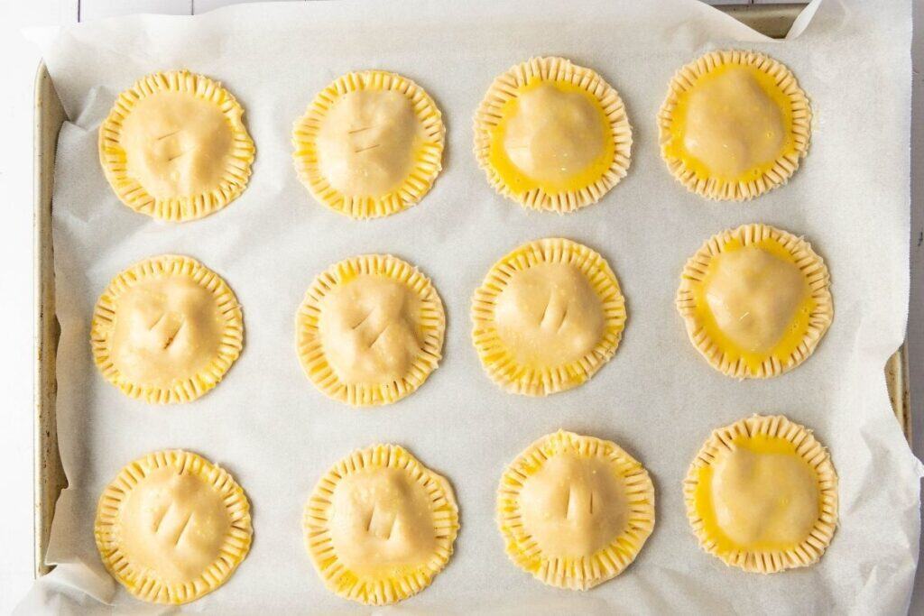 Twelve unbaked hand pies with crimped edges are arranged on a parchment-lined baking sheet, ready for the oven.