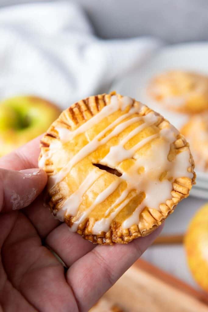 A hand holding a round, glazed pastry with crimped edges and a small slit in the center. Other pastries and a green apple are blurred in the background.