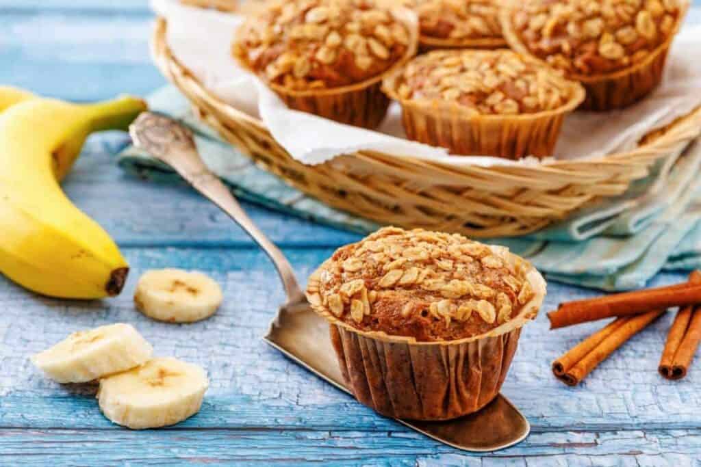 Banana muffins with oat topping on a basket and serving spatula, beside sliced bananas and cinnamon sticks on a blue wooden surface.