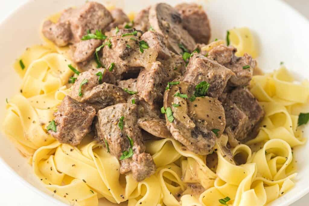 A plate of egg noodles topped with creamy beef stroganoff, mushrooms, and garnished with chopped parsley.