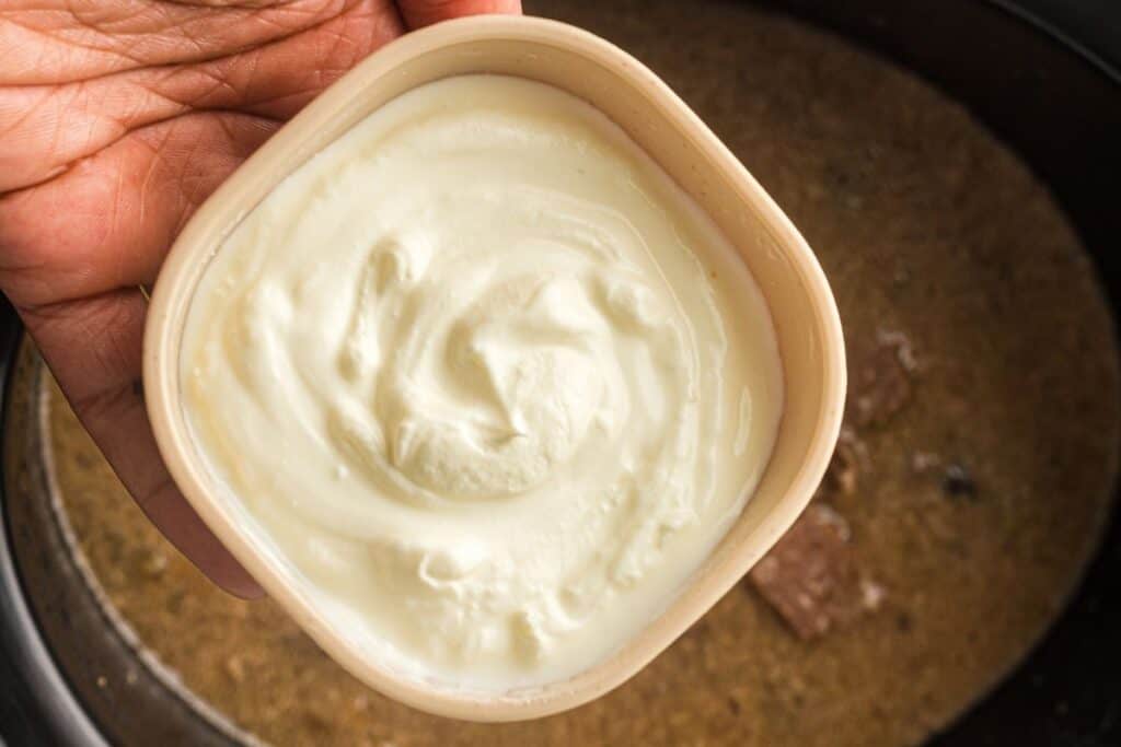 A hand holds a bowl of sour cream above a pot containing a brown, saucy dish.