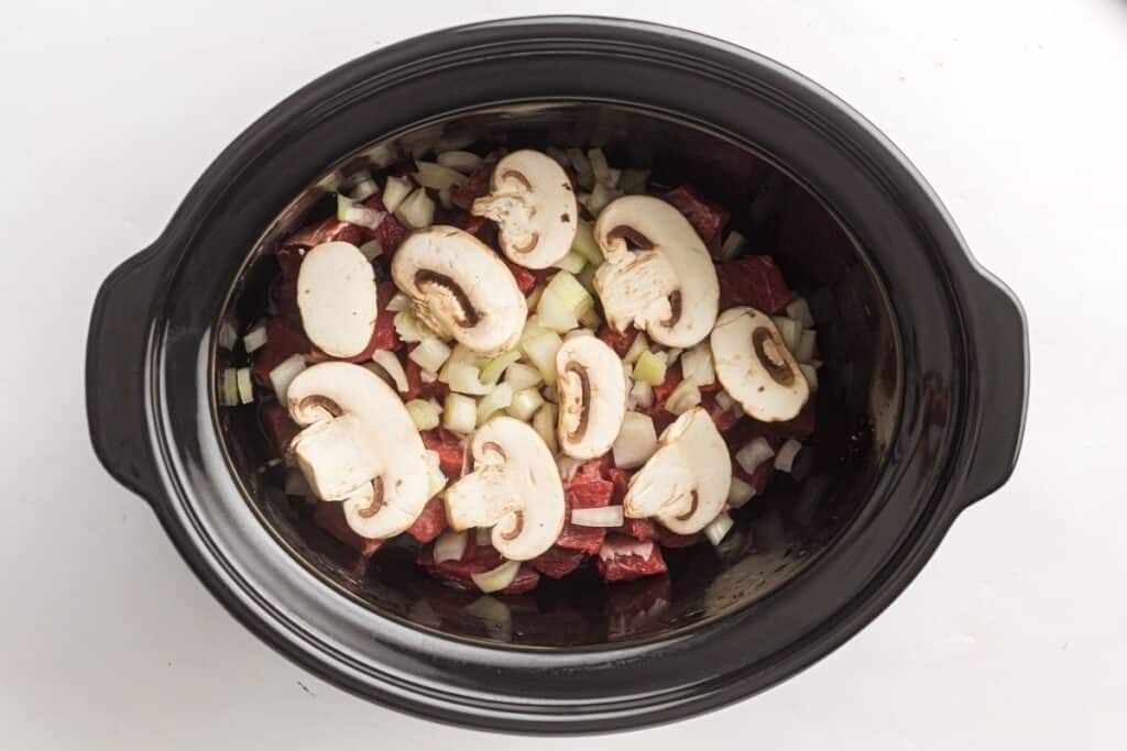 Sliced mushrooms, diced onions, and chunks of raw beef in a black slow cooker on a white surface.