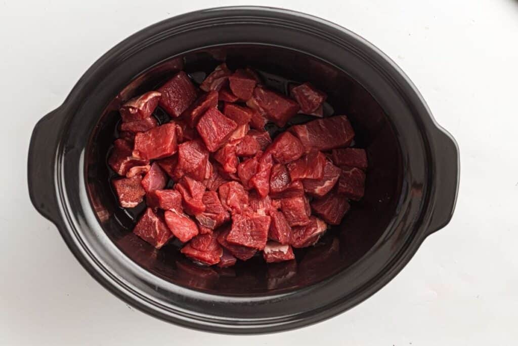 Cubed raw beef pieces placed inside a black slow cooker on a white surface.