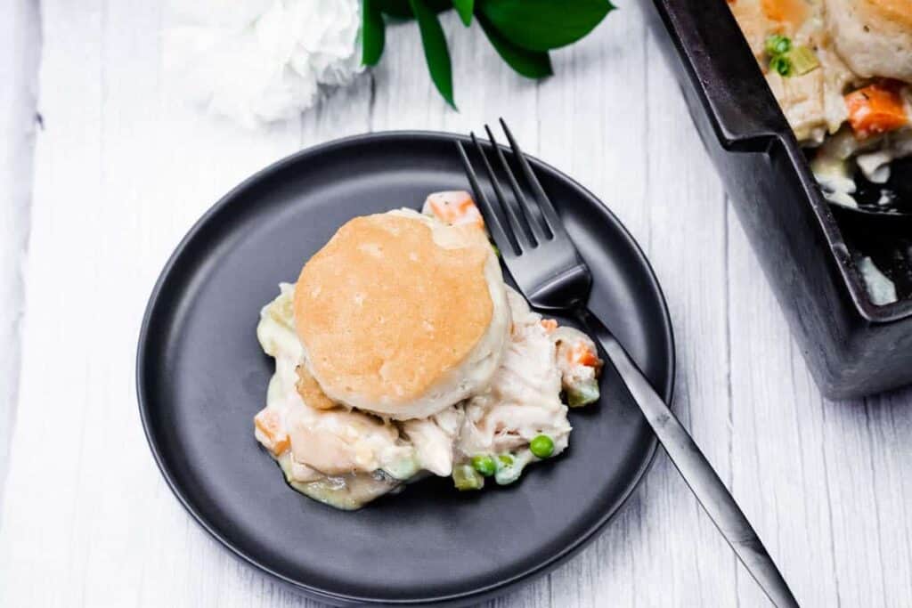 A serving of chicken pot pie topped with a biscuit, served on a black plate with a fork, next to a casserole dish on a white wooden surface.