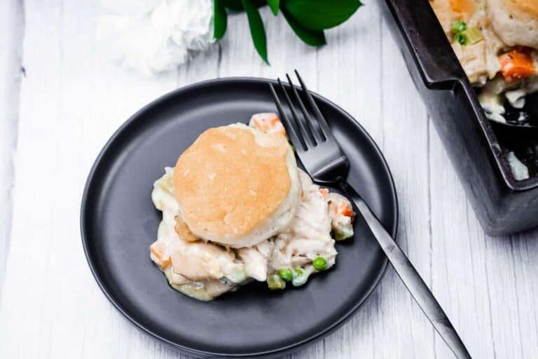 A serving of chicken pot pie topped with a biscuit, served on a black plate with a fork, next to a casserole dish on a white wooden surface.