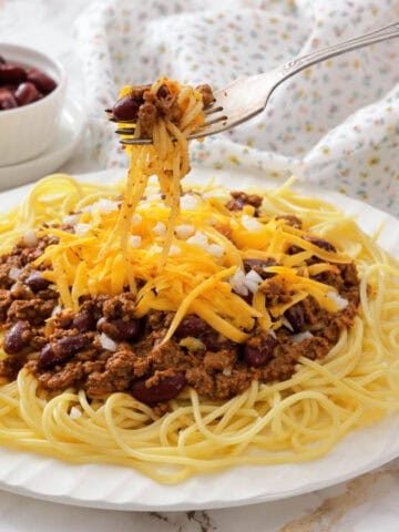 A plate of spaghetti topped with Cincinnati chili, shredded cheese, and beans, with a fork lifting a portion.