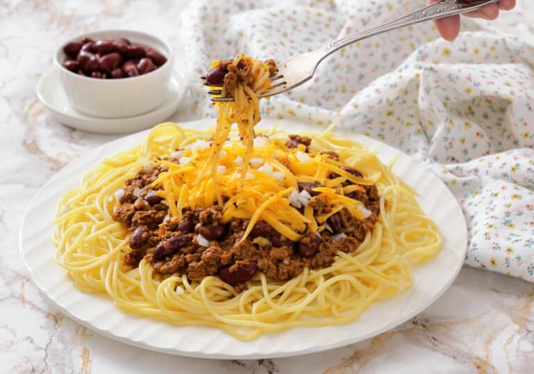 A plate of spaghetti topped with chili, kidney beans, shredded cheddar cheese, and chopped onions, with a fork lifting a portion. A small bowl of beans is in the background.